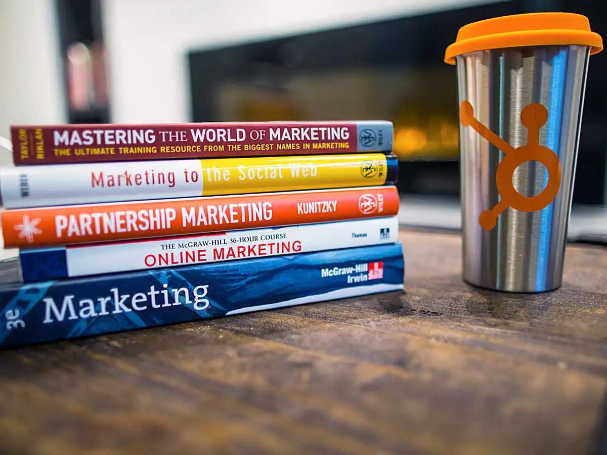 A stack of online marketing books on wooden table beside an aluminum coffee cup with orange accents.